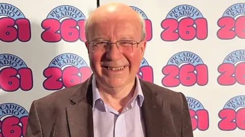 Pete Johnson Jim Hancock stands in front of the camera and smiles. He's wearing glasses and a brown suit behind a backdrop of Piccadilly Radio's logo.