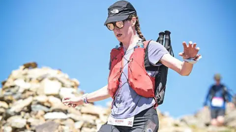 Gemma Hunter Gemma is taking part in a race and wearing a sports top, hat, sunglasses, backpack and shorts running down a countryside path with scree and piles of rocks around her.