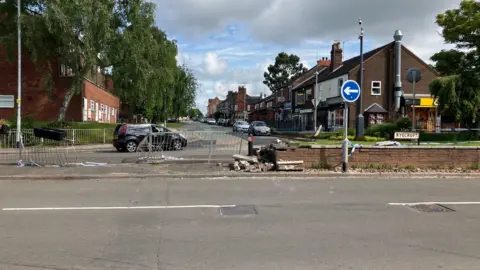 A street scene shows a damaged brick wall and temporary fencing, suggesting a recent crash. Cars and shops line the road under a cloudy sky. 