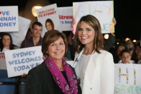 NBC / Today via Reuters Nancy Guthrie is on the left and Savannah Guthrie on the right, on the set of NBC's morning show Today. There are people in the background with 'Welcome to Sydney' banners. Nancy is smiling and wearing a purple scarf while her daughter is also smiling, wearing a white suit