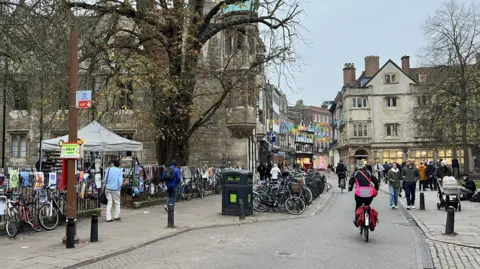 Looking down a street in Cambridge city centre. Two cyclists are riding away from the camera. Black iron railings and a row of parked bikes are to the left, and a terrace of old buildings curves to the right in the distance. Groups of pedestrians walk on the paths either side. 