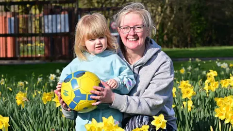 A child is holding a football and standing with their mother looking into the camera. They both have blond hair. Daffodils surround them. The woman is wearing black glasses.