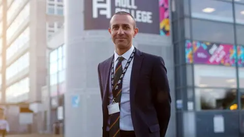 Hull College Ranjit Singh with short dark hair and wearing a dark suit, with white shirt, striped tie and lanyard stands in front of the Hull College building