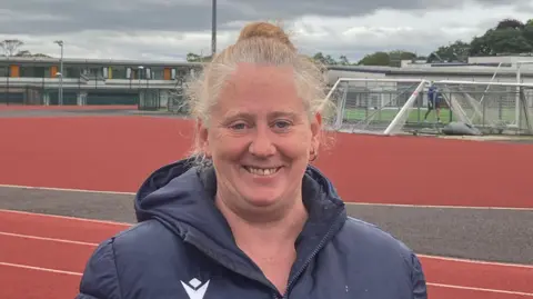 Grace Wood/BBC A woman with a blonde bun on the top of her head, wearing a navy jacket, smiles at the camera. She is standing on an athletics track