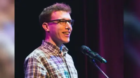 Thom Bartley Karl Knights is standing just off centre to the left, and is looking towards the right of the image. He is talking into a microphone, and is wearing a brown patterned shirt. 