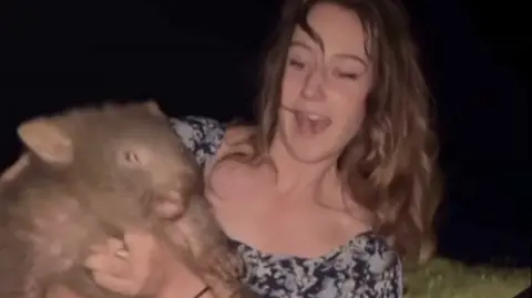 A slightly blurry screenshot of the woman holding the wombat up to the camera. She has long brown wavy hair and is wearing a floral top.