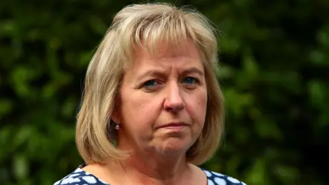 BBC Catherine O'Sullivan, who has blonde hair, looks at the camera with a sombre expression.