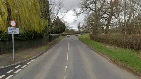 An image of a road with cars in the distance. On one side are hedges, on the left is a wall and trees with a "no access" sign.