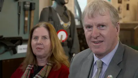 Mr Le Druillenec's grandchildren Peter and Linda smile at the camera. He is wearing a grey suit jacket and tie and she is wearing a red coat and checked scarf. Behind them is a World War Two display featuring a military uniform and a gun.