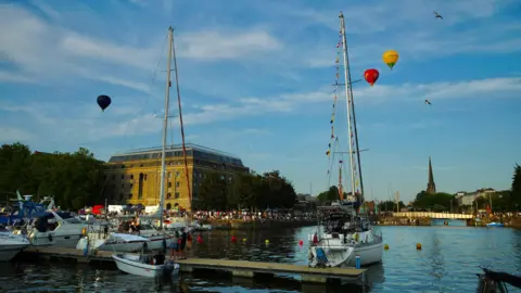 Image of Bristol's Harbour, with three hot air balloons in the air and crowds gathered near the water. 