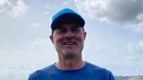 Alan Jenner wearing a blue top and blue cap. He is smiling at the camera. Behind him is a grey beach and blue sea. The sky is blue and cloudy.
