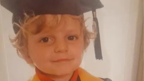 Family photo A young boy with curly blonde hair is smiling while wearing a graduation style cap and gown