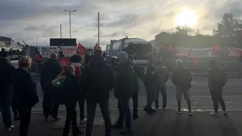 A group of people are standing on a pavement opposite a bin depot. The sun is rising in the cloudy sky. There is a large bin lorry driving out of a gate. There are red flags on the metal fences surrounding the depot exit
