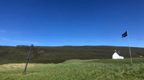 Lura The area of the range is a large hilly and grassy area. There is a white-walled house and flag.