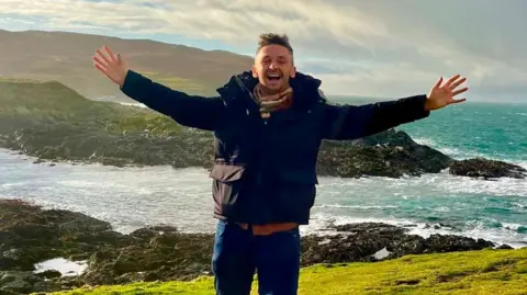 Daniel Herszberg is standing with his arms stretched wide, wearing a black coat and scarf, smiling widely. He is in front of an islet separated by a choppy sea on a bright and cloudy day.