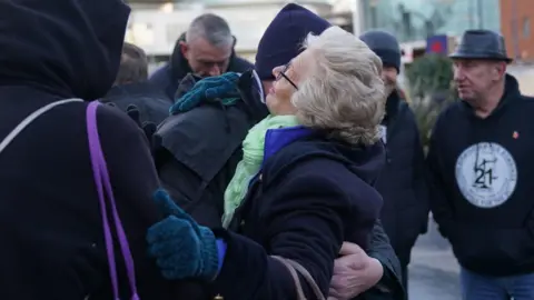 Julie Hambleton is seen hugging a fellow campaigner at a memorial service. She is wearing a coat and scarf and gloves and people around her, outdoors in the city centre, are also in hats and coats. One man has his head bowed.