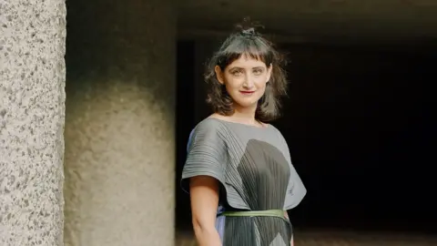 Prarthna Singh A woman wearing a grey and black dress stands outside. She has black hair tied back and a fringe. There are granite walls in the background.