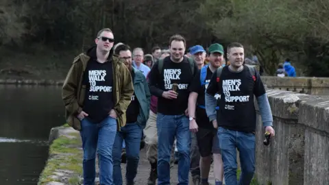 Dorothy House A group of men walking by a river. Four of the men are wearing black T-shirts which say Men's Walk to Support