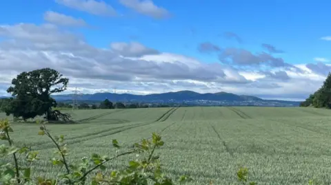 Local Democracy Reporting Service Green fields occupy roughly the bottom half of the photo. Hills are in the background, beneath blue skies and mainly grey clouds.