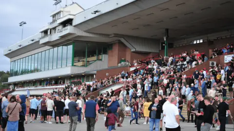 Crowds are in the stands and on ground level at the racecourse. Many are wearing T-shirts. The stadium can be seen in the background and it is a glass and metal building with large windows looking onto the course.
