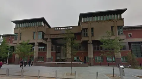 Exterior of modern red-brick and brown court building, with columns on its facade. A group of people is standing in front of the building to one side, while two trees stand in the foreground.