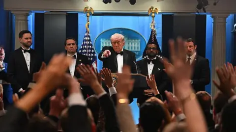 Anadolu via Getty Images Trump, wearing a bow tie and tuxedo, points to a reporter in a room full of raised hands trying to get his attention.