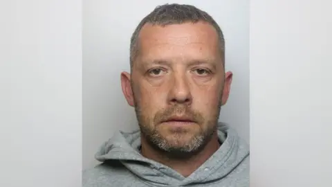 National Crime Agency A custody shot of Kevin Newson. He is wearing a grey hoodie and is staring into the camera. He has short brown hair, flecked with grey, a short beard of the same colour.