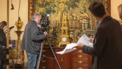 BBC Studios/ Anna Gordon A film crew inside the Abbey looking at a detailed tapestry of birds and trees. A man with a camera on a tripod is stood in front of a wooden set of draws to film the art. A woman with a boom microphone stand behind him.