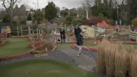 Children and parents playing at Mr Mulligan's Dino Golf in Tonbridge, Kent. Three people holding golf clubs can be seen in the middle of the image. 