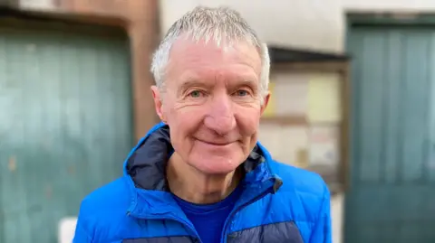 An image of a man with grey hair, wearing a blue coat