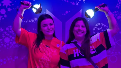 Stephanie Prado, left, wearing a red shirt, poses with her friend while holding an official BTS light stick in her right hand