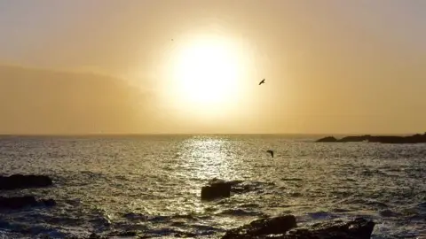 Mousehold Pat A large, glowing sun in an orange sky above ripply waves off Mousehole in Cornwall.
