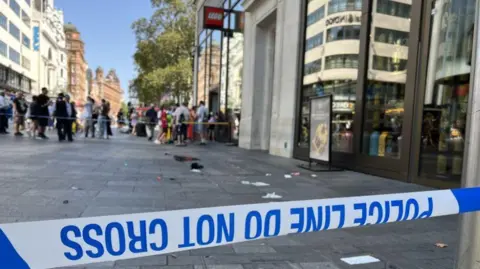 BBC Blue and white police tape outside the stabbing scene with pieces of white paper and fabric on the ground and a crowd