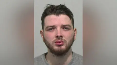 Mugshot of Stephen Scott. He has short dark hair swept back up on his head and a gingery straggly beard. His face is red and his thick lips appear to be in a slight pout.