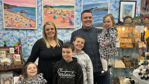 Rachel Rasmussen Rachel in a black top with her husband in a navy green checked shirt. He is holding a toddler girl and their three other children stand in front of the couple - two boys and one girl. Some of Rachel's paintings of the beach are displayed on the wall behind them