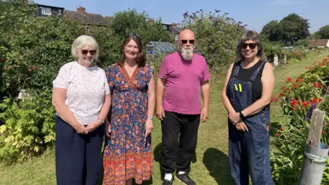 Plot holders Ann, Lisa, Mick and Catharine standing together in Brookfield Allotment on a grass path in front of flowers and shrubs.