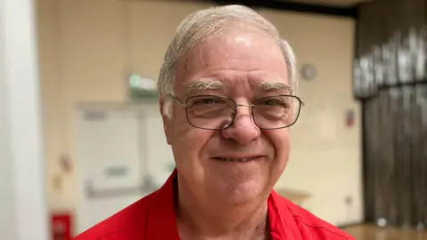 The President of the IDOLS drama group Ian Phillips is smiling for the photo. He is wearing glasses and a red shirt.