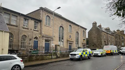BBC Large old stone building with large arched windows. Police vehicles are parked on the road outside and police tape is strung along the railings.