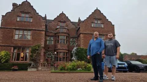Emporium Productions Two men, one in a blue top and trousers and another in a grey T-shirt and blue jeans down to the knee, are standing a fair way from the camera. They are standing outside a large home - the brown building has three storeys and, photographed outside from this side, could have three rooms on each storey on this side.