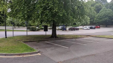 Keswick Lakeside Car park. Four empty spaces sit underneath a tree. Several other cars are parked in the background.