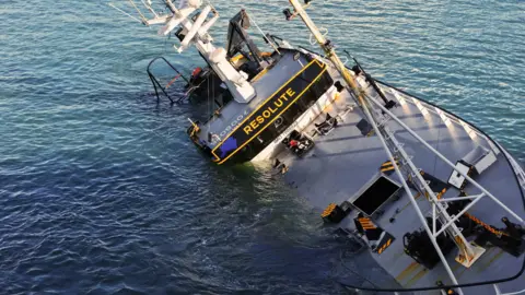 JC Explores Close up drone shot shows the large navy and orange vessel sunk in the sea leaning on one side. The deck of the boat can be seen.