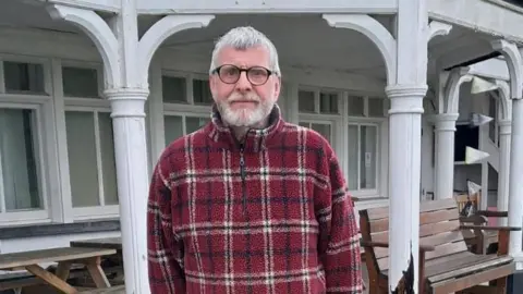 Trowbridge Cricket Club Steve Wordley wears a red tartan fleece and glasses, looking into the camera while standing outside the building.