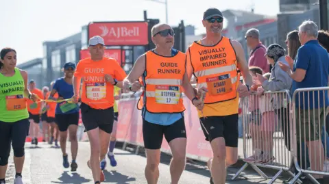 AJ Bell People are running on a road with people cheering them on. The people are wearing orange high-vis shirts saying they are guide runners or visually impaired runners. The sun is shining and everyone is laughing.