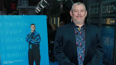 Getty Images John Davidson stood in front of a film poster for I Swear