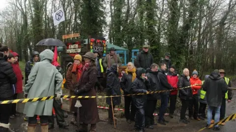 A group of campaigners stood in a field, with a line of trees behind them. They are stood behind yellow and black tape.