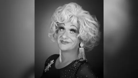 A black and white photo of Jason Sutton looking at the camera and smiling, wearing long fake eyelashes with a blond wig and dangling earring, and a black top with sparkles on it.