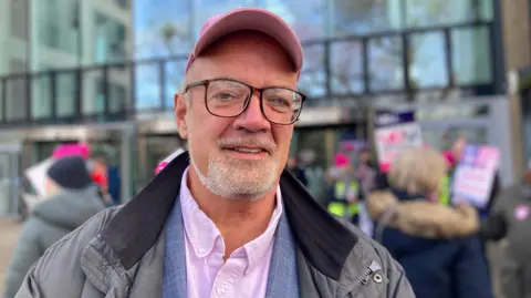 BBC/Briony Leyland A man standing alongside a picket line outside a large building. He is wearing a pink cap and shirt with a grey jacket. He has black glasses and a short grey beard. People behind are photographed out of focus and are holding pink, white and purple signs.