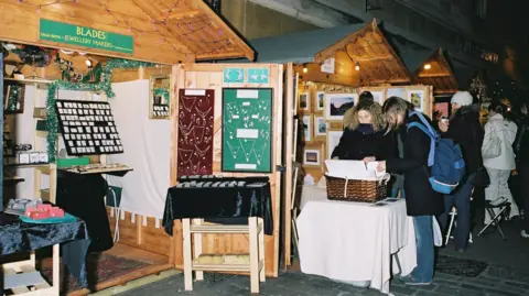 B&NES Council A line of chalets with the nearest one selling jewellery. Two women are looking at a basket of prints at another stall selling paintings.