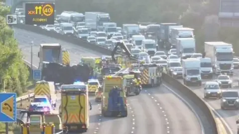 One carriageway of a motorway is closed with emergency vehicles parked up. There is a large overturned vehicle surrounded by emergency services. 