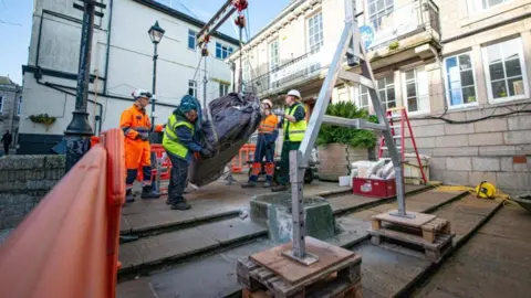 Four people in high-vis jackets are outside a building and are helping to manoeuvre the wrapped sculpture which is being suspended by a small crane.